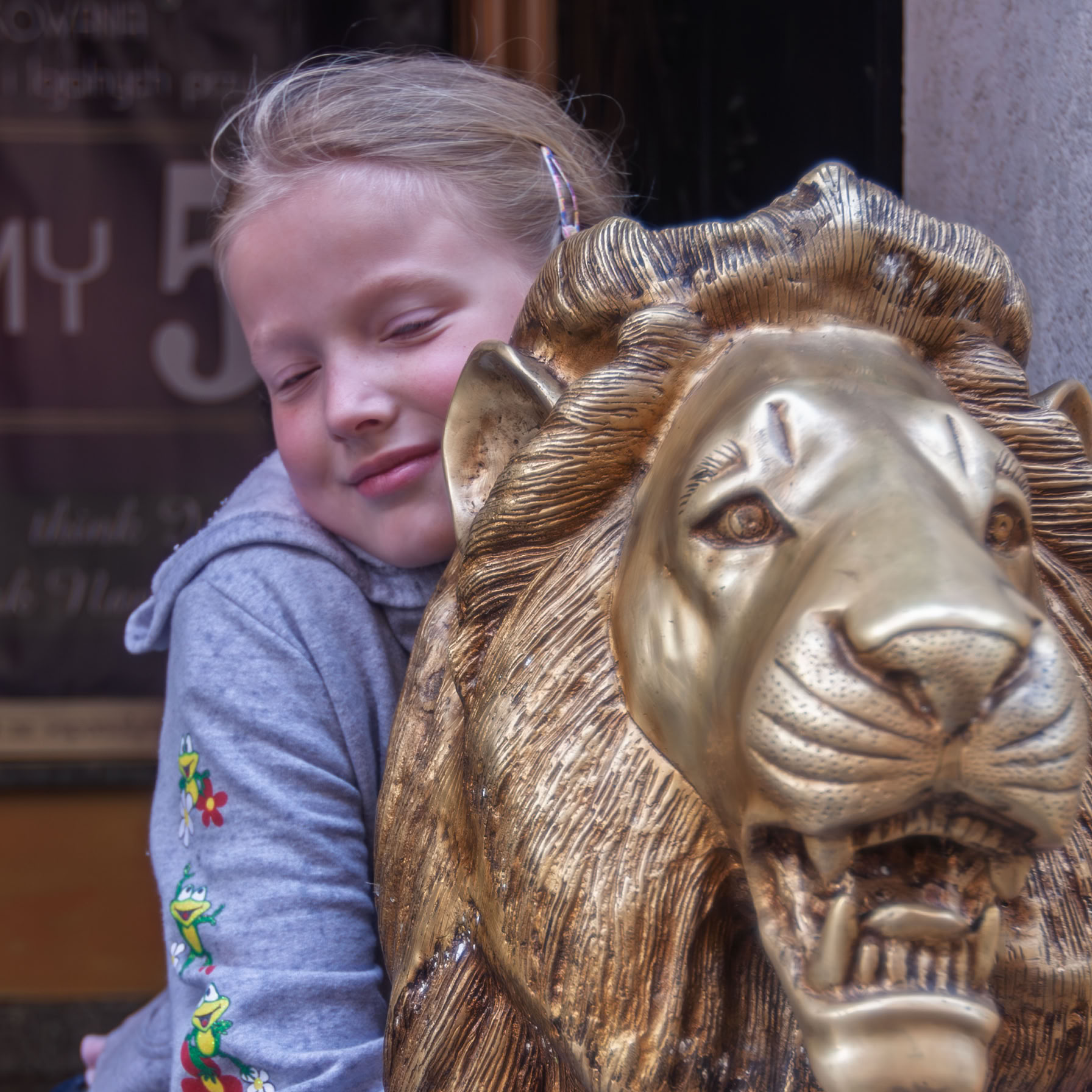 Mała dziewczynka o blond włosach usiadła na posągu lwa i go mocno tuli do siebie.