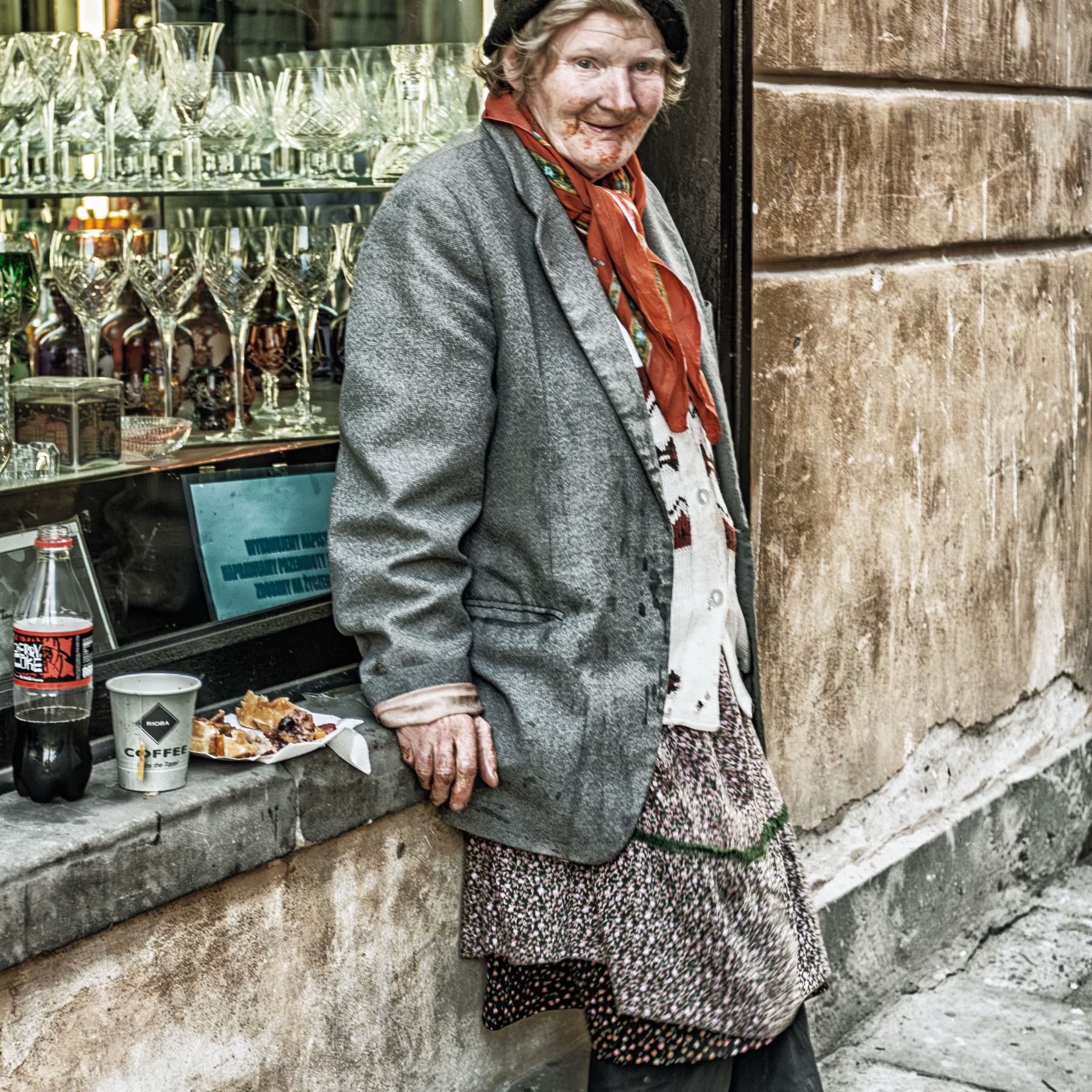 Piękna starość. Starsza kobieta oparta o witrynę sklepu z Kiliszkami.