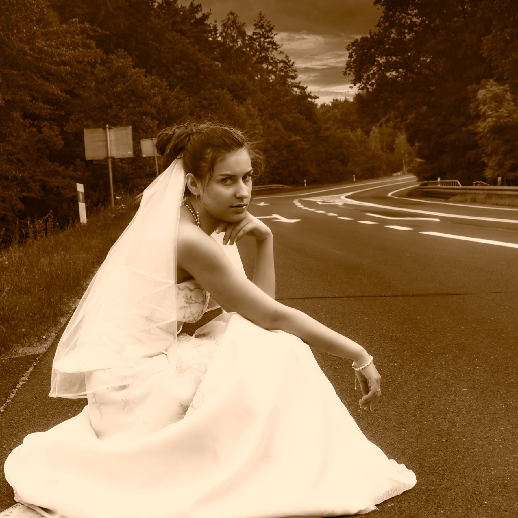 Młoda dziewczyna w sukni ślubnej przykucnęła na drodze, czeka na okazję auto-stopu.